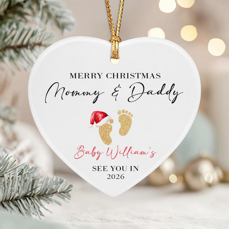 A white heart-shaped ceramic ornament with a gold hanging string. It features the text MERRY CHRISTMAS Mommy & Daddy, Baby William's, and SEE YOU IN 2026, along with two gold glitter baby footprints, one wearing a red Santa hat.