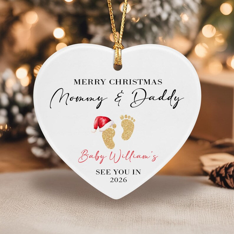 A white heart-shaped ceramic ornament hanging by a gold cord. It features the text MERRY CHRISTMAS Mommy & Daddy, Baby William's, and SEE YOU IN 2026. Two gold glitter baby footprints are centered, one wearing a red Santa hat.