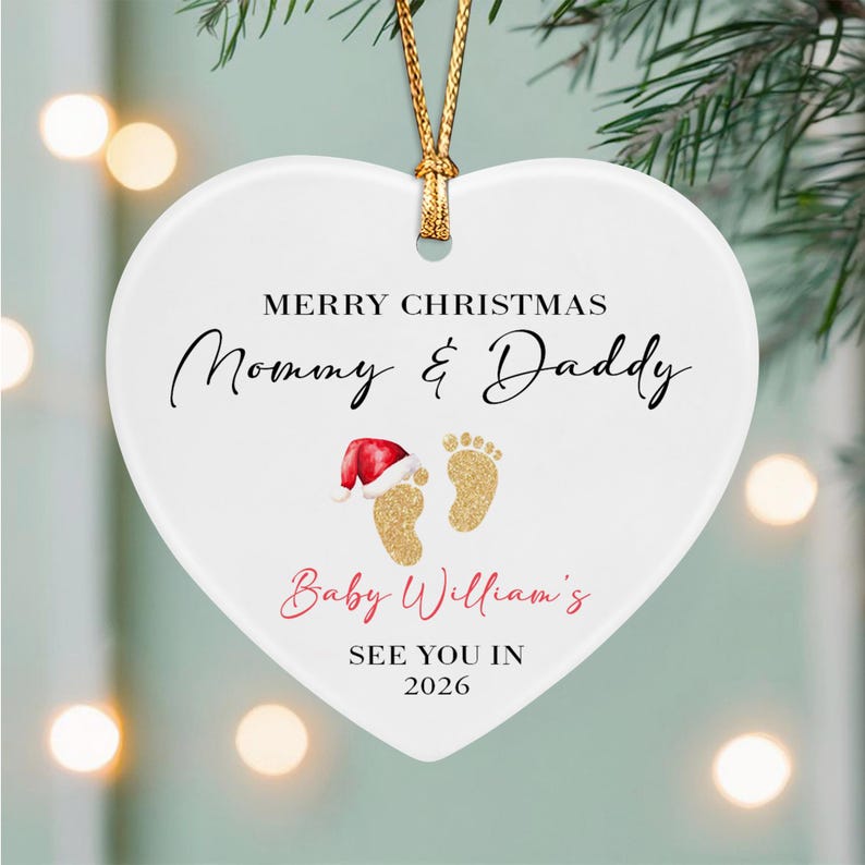 A white heart-shaped ceramic ornament hanging from a gold string. It features the text Merry Christmas Mommy & Daddy, Baby William's, and See You In 2026, with a graphic of two gold baby footprints, one wearing a red Santa hat.