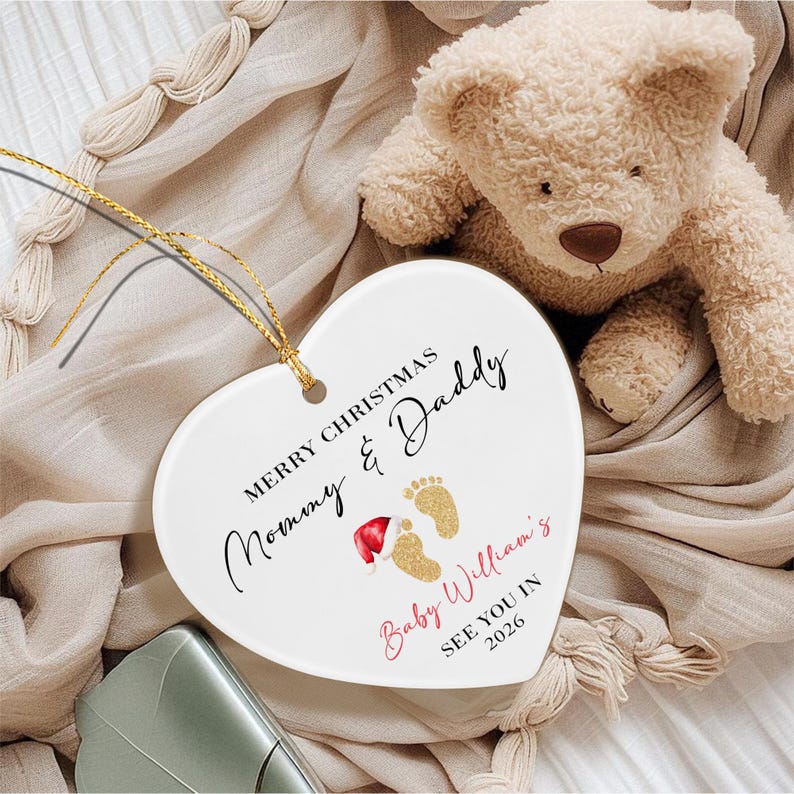 A white heart-shaped ceramic ornament with a gold string. The text reads MERRY CHRISTMAS MOMMY & DADDY, Baby William's, SEE YOU IN 2026. It features two gold glitter baby footprints, one wearing a small red and white Santa hat.