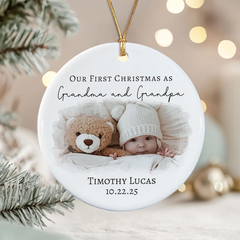 A round white ceramic ornament featuring a gold hanging string. The text reads Our First Christmas as Grandma and Grandpa, Timothy Lucas, and 10.22.25.