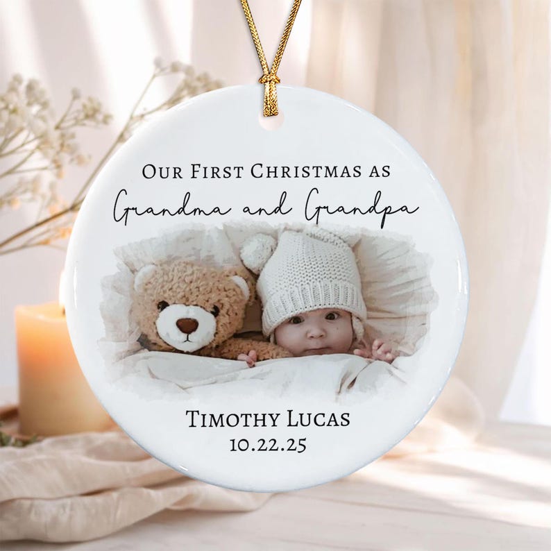 A round white ceramic ornament hanging from a gold cord. The text reads Our First Christmas as Grandma and Grandpa, Timothy Lucas, and 10.22.25. A photograph shows a baby in a white knit hat next to a brown teddy bear.