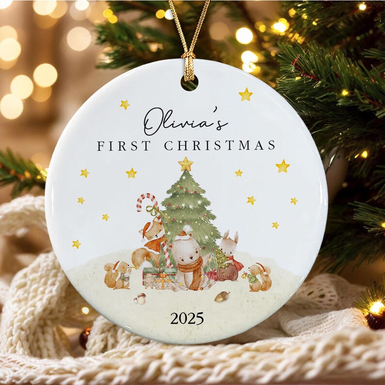 A round white ceramic ornament featuring the text Olivia's FIRST CHRISTMAS and 2025. The design includes a green Christmas tree, a fox, a bear, and a rabbit wearing scarves, surrounded by yellow stars and a gold hanging cord.