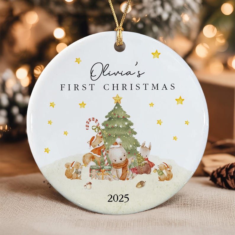 A round white ceramic Christmas ornament featuring a gold string hanger. The text reads Olivia's FIRST CHRISTMAS 2025. The illustration shows a green Christmas tree, a fox, a bear, and squirrels with gifts and stars.