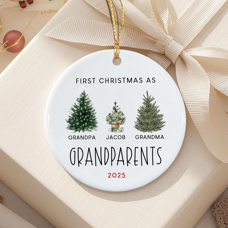 A round white ceramic ornament with a gold hanging string. The text reads FIRST CHRISTMAS AS GRANDPARENTS, with three illustrated evergreen trees labeled GRANDPA, JACOB, and GRANDMA, and the red printed year 2025 at the bottom.