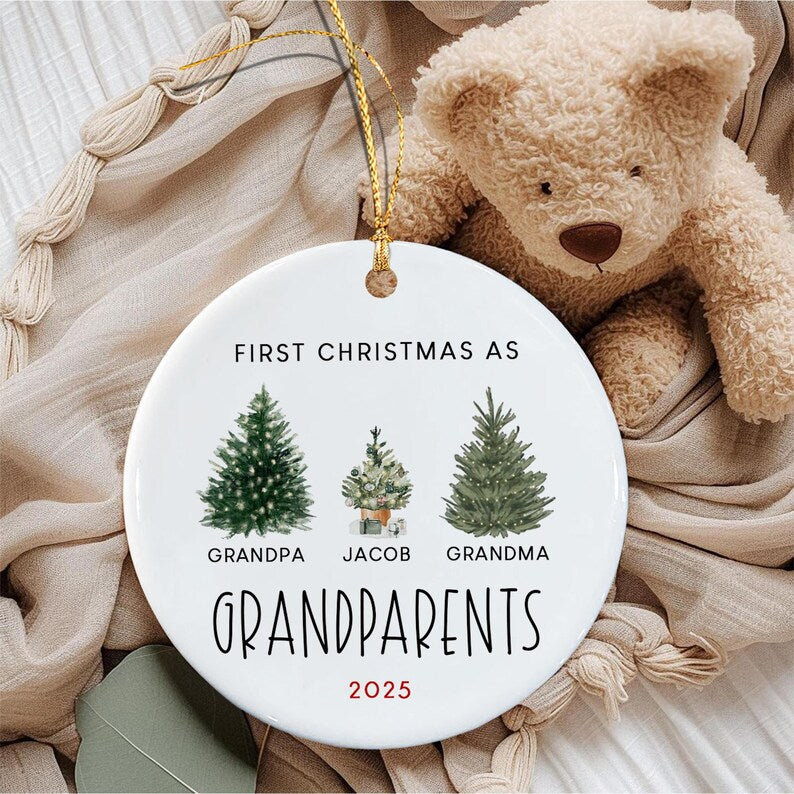 A round white ceramic ornament with a gold hanging string. The text reads FIRST CHRISTMAS AS GRANDPARENTS 2025. It features three illustrated evergreen trees with the names GRANDPA, JACOB, and GRANDMA printed below each tree.