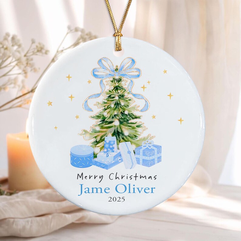 A round white ceramic ornament featuring a watercolor illustration of a green Christmas tree topped with a light blue bow, surrounded by blue gift boxes and gold stars. The text reads Merry Christmas, Jame Oliver, and 2025.