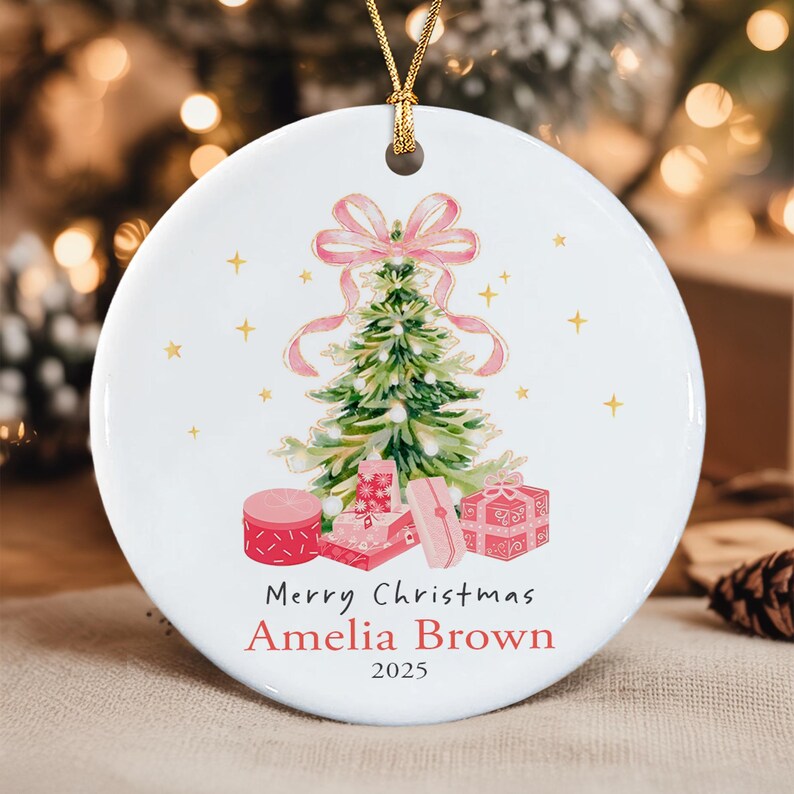 A round white ceramic ornament featuring a watercolor illustration of a green Christmas tree topped with a pink bow, surrounded by pink gift boxes.