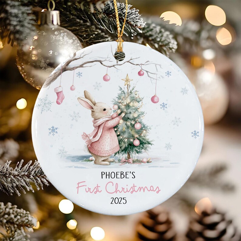 A round white ceramic ornament featuring a watercolor illustration of a rabbit in a pink sweater decorating a small Christmas tree.