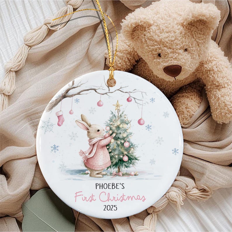 A round white ceramic ornament featuring a brown rabbit wearing a pink sweater decorating a green Christmas tree. The design includes pink ornaments, snowflakes, and the text PHOEBE'S First Christmas 2025.
