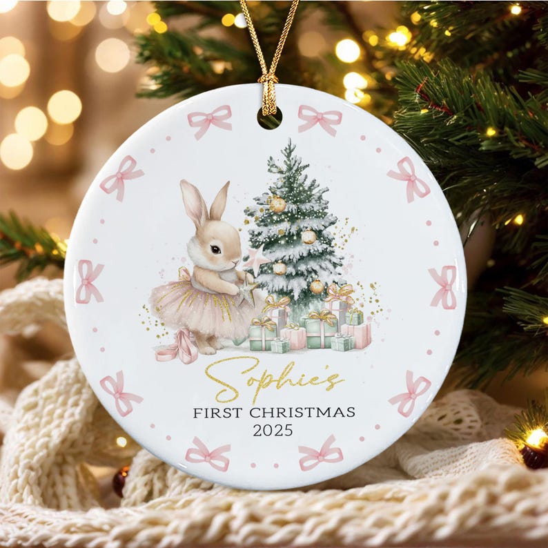 A round white ceramic ornament featuring a bunny in a tutu standing next to a decorated Christmas tree and gifts. The ornament is bordered by small pink bows and includes the gold text Sophie's FIRST CHRISTMAS 2025.