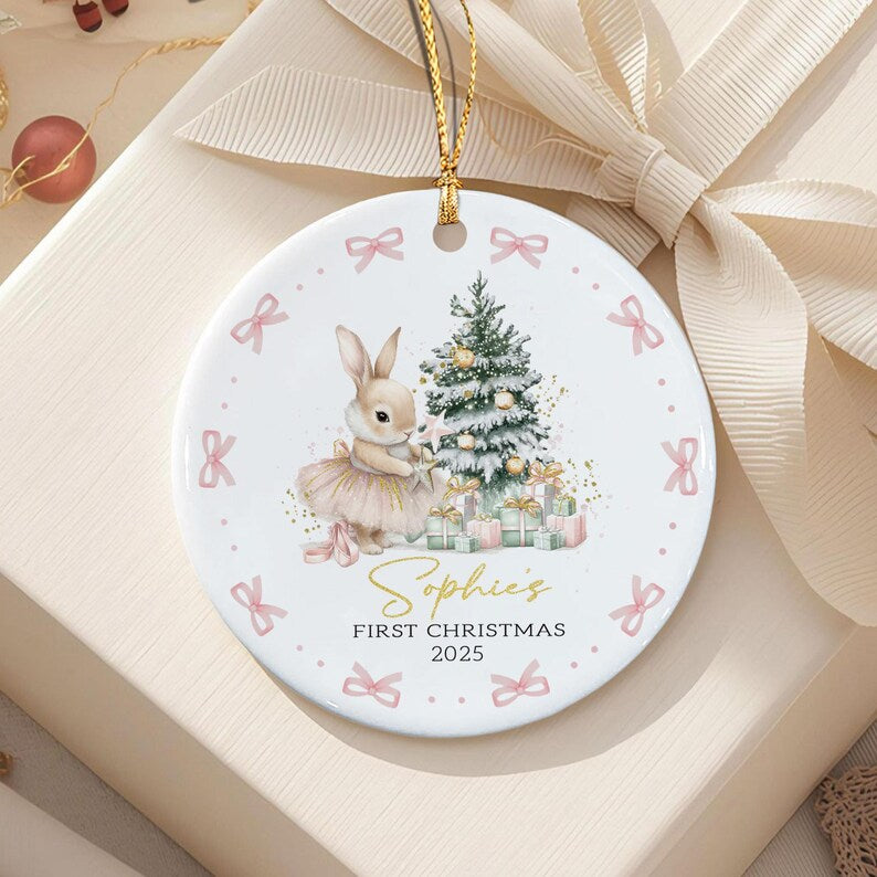 A round white ceramic ornament featuring a rabbit in a tutu standing next to a decorated Christmas tree and gifts. The text reads Sophie's FIRST CHRISTMAS 2025. The border is decorated with small pink bows and gold dots.