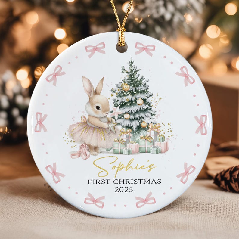 A round white ceramic ornament featuring a bunny in a tutu standing by a snow-covered Christmas tree with gifts. The text reads Sophie's FIRST CHRISTMAS 2025 in gold and black. The border is decorated with small pink bows.