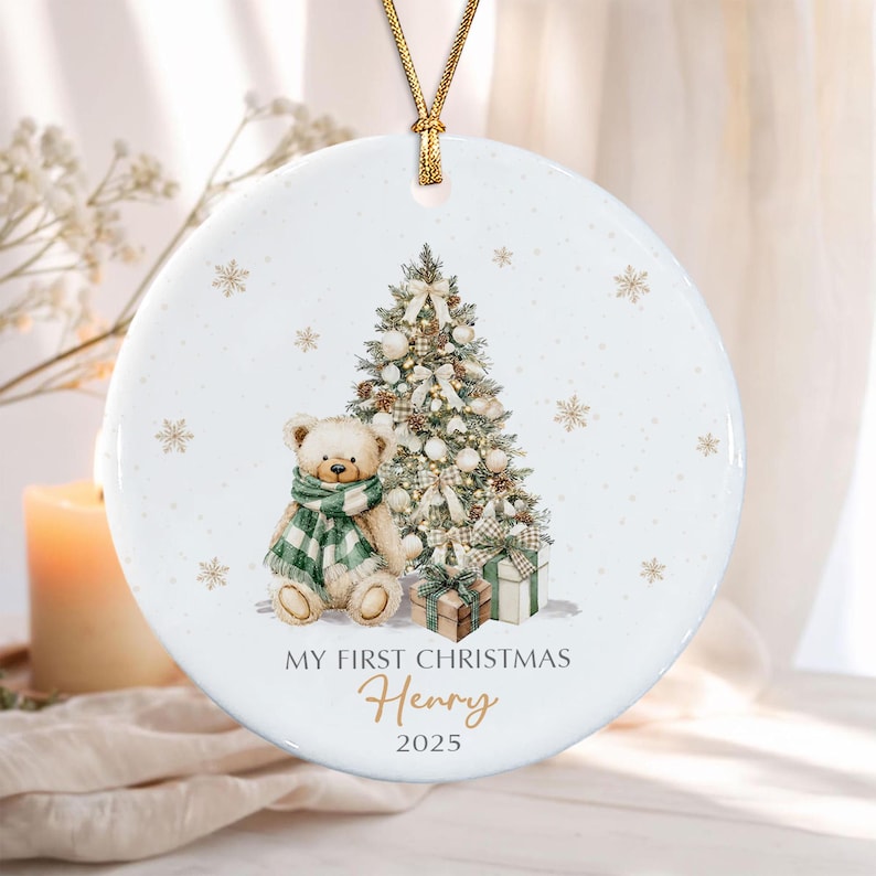 A round white ceramic Christmas ornament hanging from a gold cord. The design features a teddy bear wearing a green and white plaid scarf, a decorated Christmas tree, and gift boxes. Text reads: MY FIRST CHRISTMAS Henry 2025.