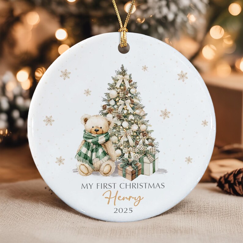 A round white ceramic ornament featuring a teddy bear wearing a green and white plaid scarf, a decorated Christmas tree, and wrapped gifts. The text reads MY FIRST CHRISTMAS, the name Henry, and the year 2025 in gold and black.