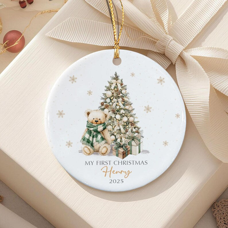 A round white ceramic ornament featuring a teddy bear wearing a green and white plaid scarf, a decorated Christmas tree, and wrapped gifts. The text reads MY FIRST CHRISTMAS, the name Henry, and the year 2025 in gold and black.