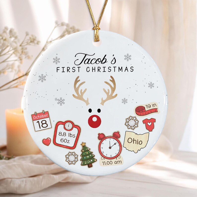 A round white ceramic Christmas ornament hanging by a gold cord. It features the text 'Tacob's FIRST CHRISTMAS' above a reindeer face.