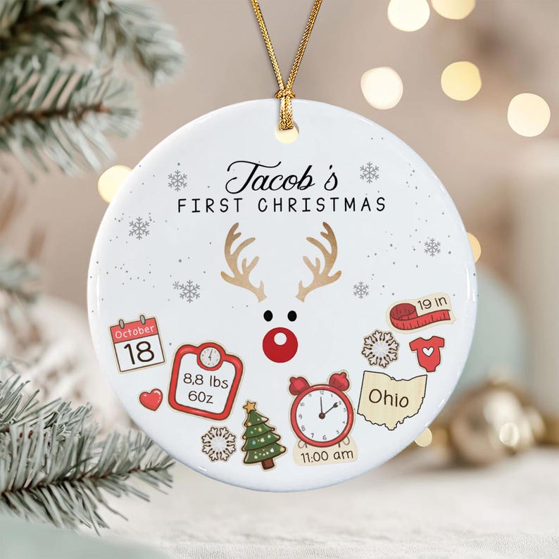 A white round ceramic ornament with a gold string featuring the text 'Jacob's FIRST CHRISTMAS' and a reindeer graphic.