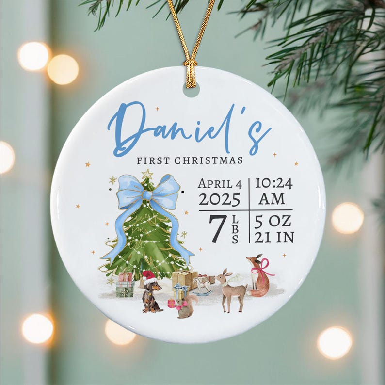 A round white ceramic ornament with a gold string featuring the text Daniel's FIRST CHRISTMAS, a green Christmas tree with a blue bow, a dachshund, a deer, a fox, and birth details: April 4 2025, 10:24 AM, 7 LBS 5 OZ, 21 IN.