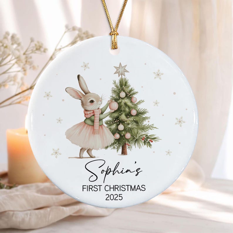 A round white ceramic ornament featuring a watercolor illustration of a rabbit in a pink tutu and scarf decorating a Christmas tree. The text reads Sophia's FIRST CHRISTMAS 2025. The ornament hangs from a gold cord.