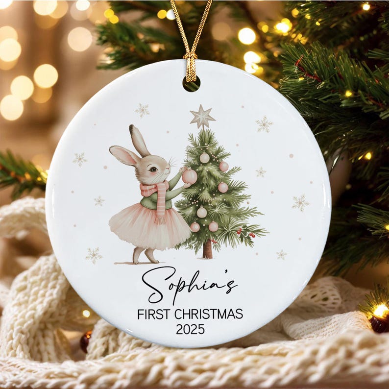 A round white ceramic ornament featuring a rabbit wearing a pink skirt and scarf, decorating a green Christmas tree with pink baubles. The text reads Sophia's FIRST CHRISTMAS 2025.