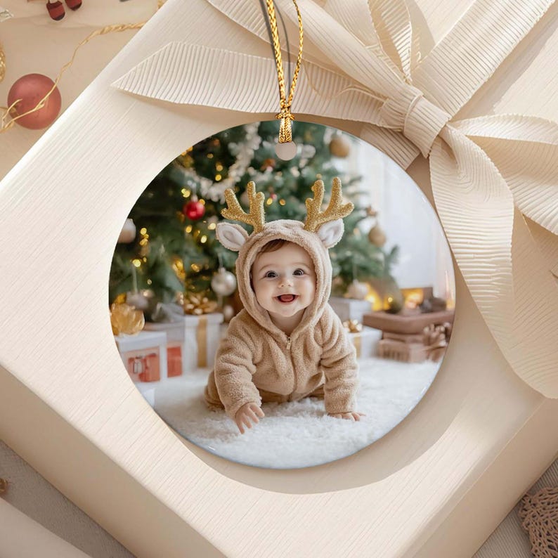 A round ceramic ornament featuring a photograph of a baby wearing a tan hooded jacket with gold glitter reindeer antlers. The ornament is suspended by a gold string and is displayed against a white gift box with a large ribbon.