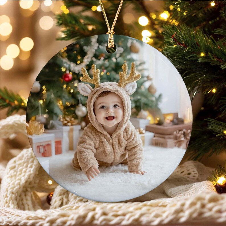 A round ceramic Christmas ornament featuring a photograph of a baby wearing a tan hooded onesie with gold glittery reindeer antlers.