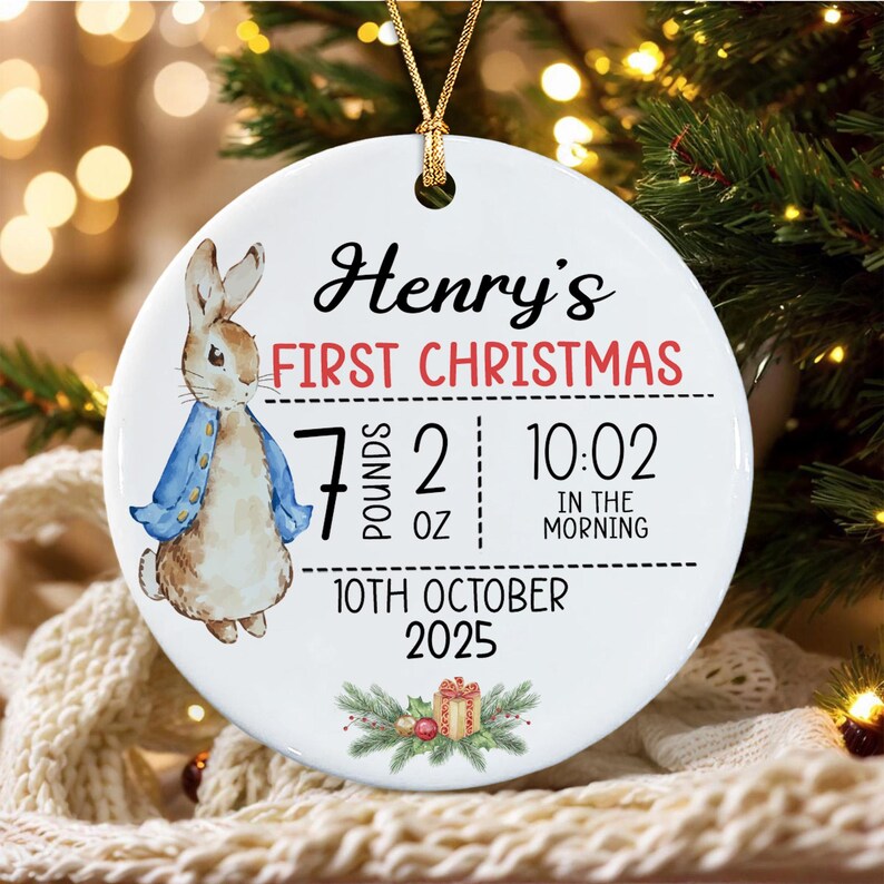 A round white ceramic ornament featuring a watercolor illustration of a rabbit in a blue jacket. Text reads: Henry's FIRST CHRISTMAS, 7 POUNDS 2 OZ, 10:02 IN THE MORNING, 10TH OCTOBER 2025.
