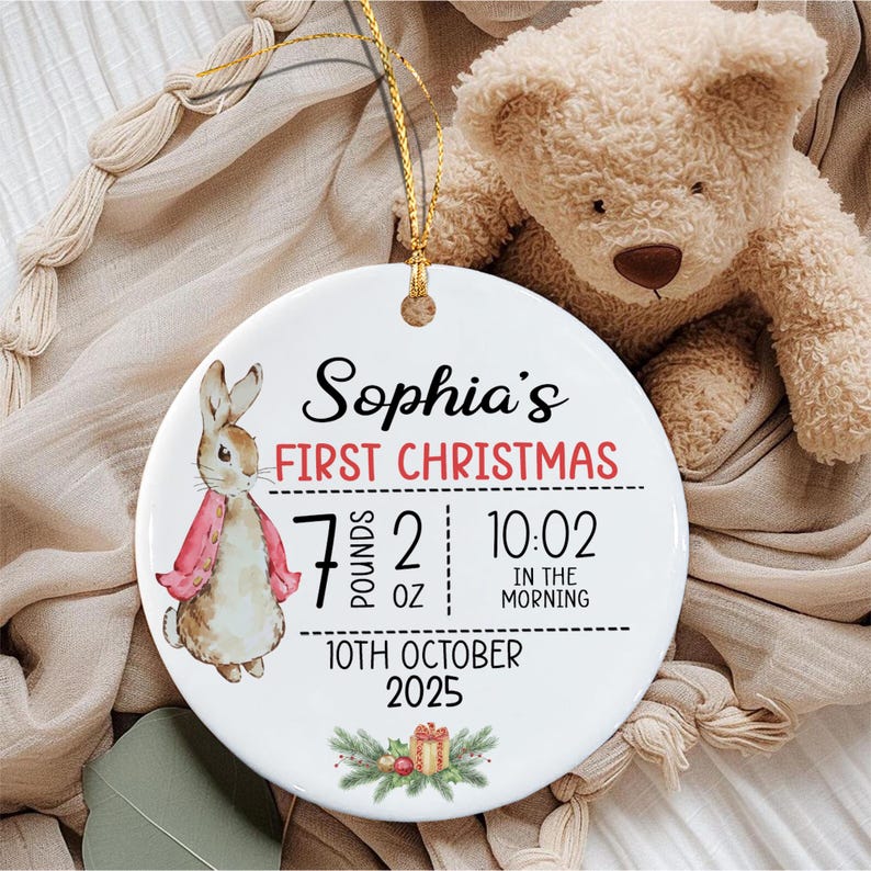 A round white ceramic ornament featuring a watercolor illustration of a rabbit in a red jacket. Text reads: Sophia's FIRST CHRISTMAS, 7 POUNDS 2 OZ, 10:02 IN THE MORNING, 10TH OCTOBER 2025.