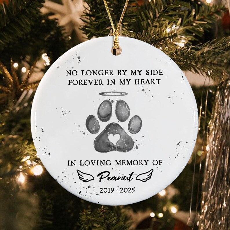 A round white ceramic ornament featuring a gray paw print with a halo and a heart cutout. The text reads: NO LONGER BY MY SIDE FOREVER IN MY HEART, IN LOVING MEMORY OF Peanut, and 2019-2025. It hangs from a gold string.