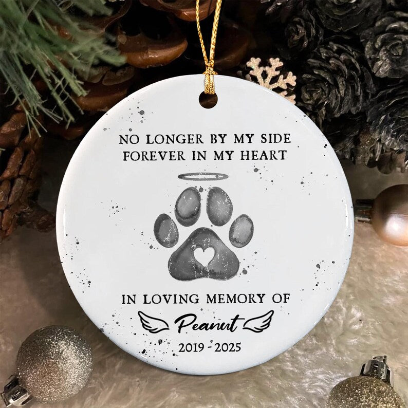 A round white ceramic ornament featuring a gray paw print with a halo and a small heart cutout. The text reads: NO LONGER BY MY SIDE FOREVER IN MY HEART, IN LOVING MEMORY OF Peanut, and 2019-2025, accented with small wings.