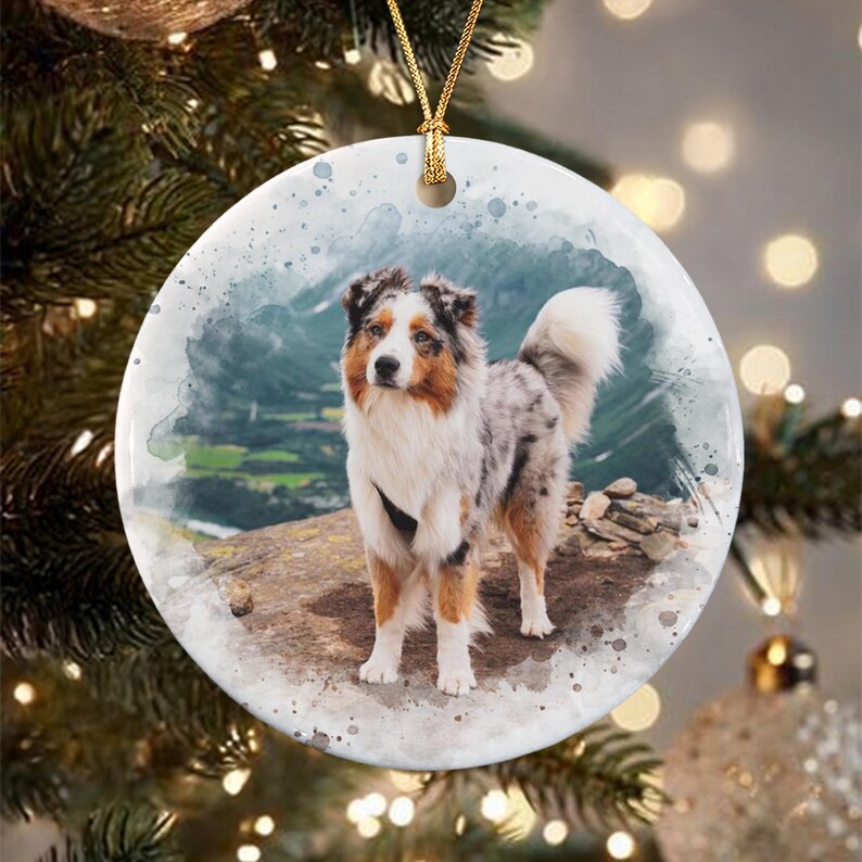 A round white ceramic ornament featuring a watercolor-style illustration of an Australian Shepherd dog standing on a rocky cliff overlooking a valley. The ornament is suspended by a gold string against a blurred tree background.
