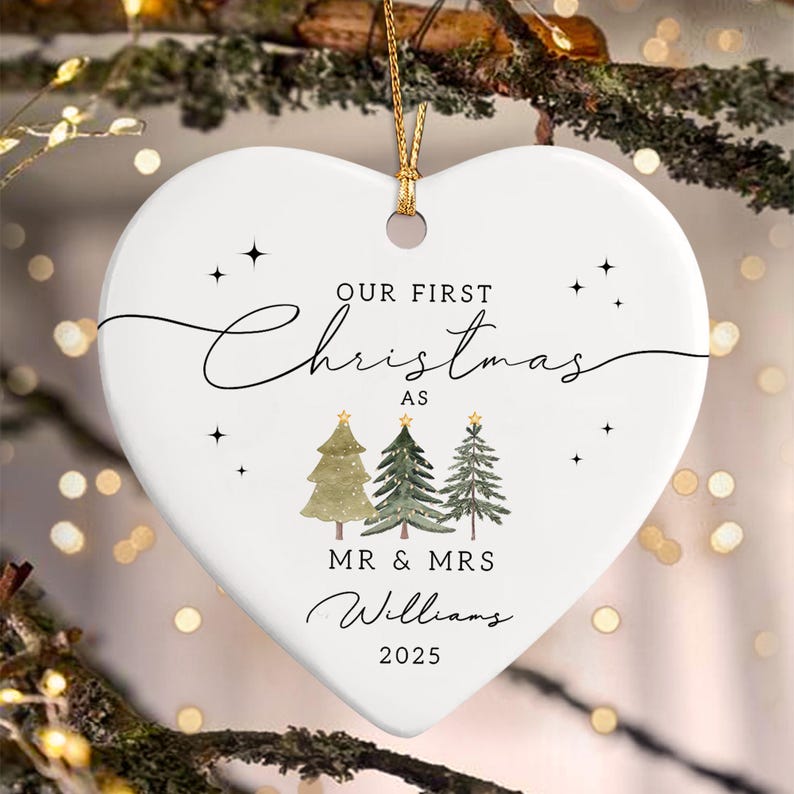 White heart-shaped Christmas ornament with gold hanging cord. The ornament features the text "OUR FIRST Christmas AS" above three illustrated Christmas trees. Below the trees is "MR & MRS Williams 2025".