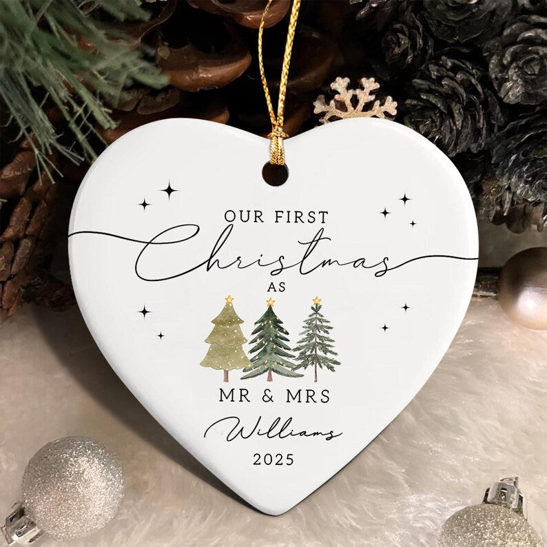 White heart-shaped Christmas ornament with gold string. The ornament features the text "Our First Christmas" and "MR & MRS Williams 2025". Three green Christmas trees are below the text.