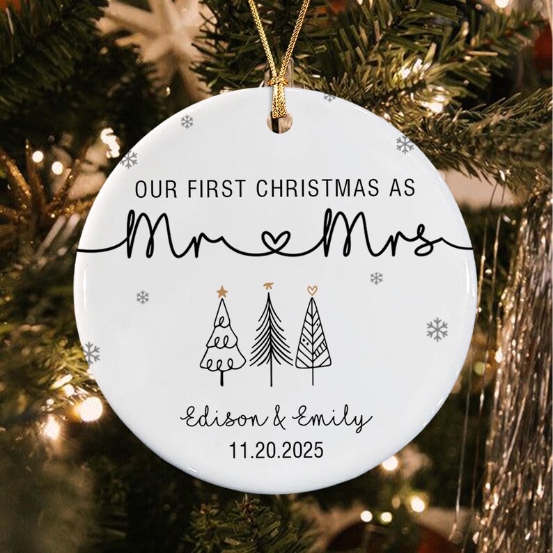 A round white ceramic ornament featuring the black text "OUR FIRST CHRISTMAS AS Mr & Mrs" with a heart symbol, three stylized black line-art Christmas trees, and the names "Edison & Emily" with the date "11.20.2025" in black.