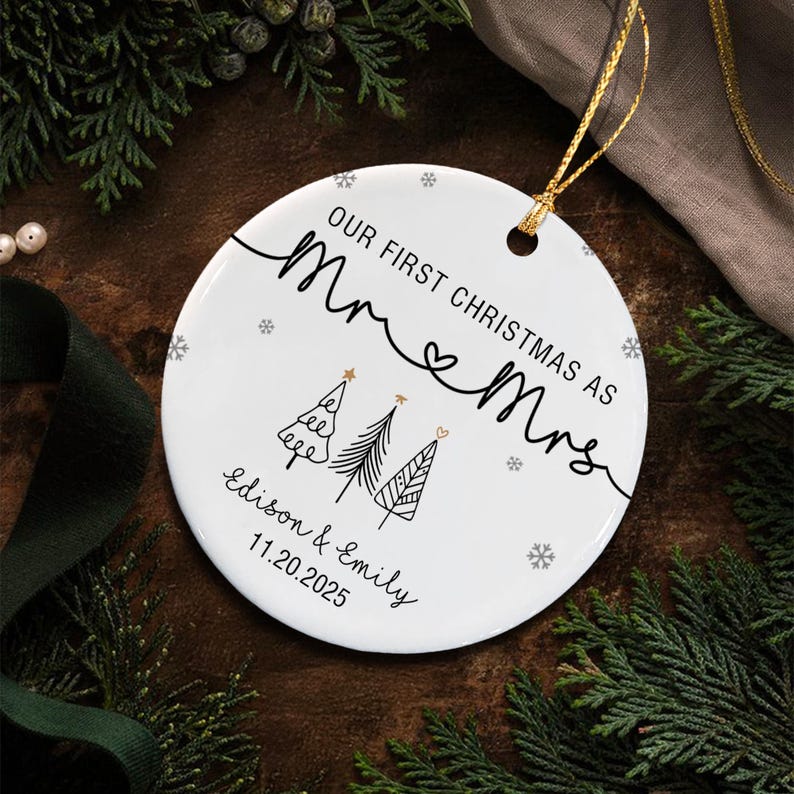 A round white ceramic ornament featuring the black text Our First Christmas As Mr & Mrs, three stylized line-art Christmas trees, and the names Edison & Emily with the date 11.20.2025. It includes a gold hanging string.