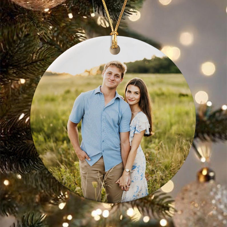 A round ceramic ornament featuring a photograph of two people standing in a grassy field. The person on the left wears a light blue button-down shirt and tan pants, while the person on the right wears a white floral print dress.