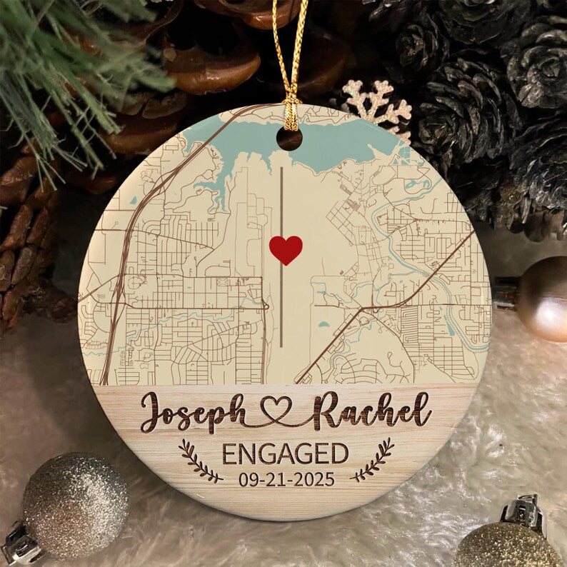 A round ceramic ornament featuring a cream-colored map with a red heart, suspended by a gold string. The lower section has a wood-grain pattern with the text Joseph & Rachel, ENGAGED, and the date 09-21-2025 printed in dark brown.