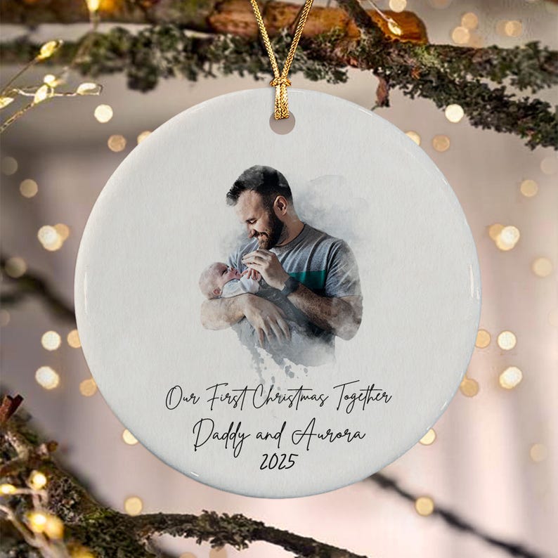 A round white ceramic ornament featuring a watercolor-style illustration of a person holding a baby. Below the image, the text reads: Our First Christmas Together, Daddy and Aurora, 2025. A gold string is attached to the top.