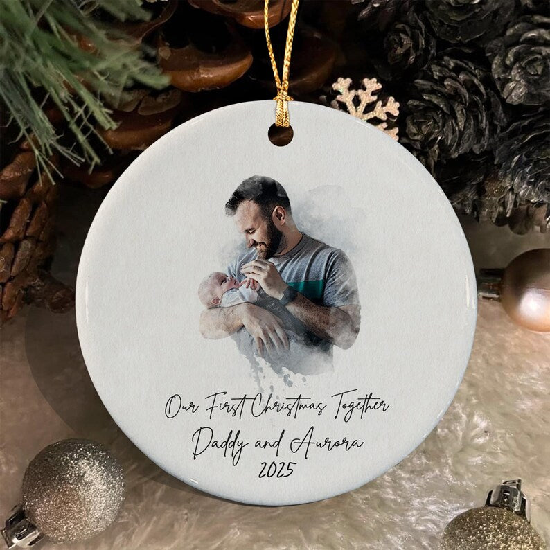A round white ceramic ornament featuring a printed watercolor-style image of a parent holding a baby. Below the image, black cursive text reads: Our First Christmas Together, Daddy and Aurora, 2025. A gold string is attached.
