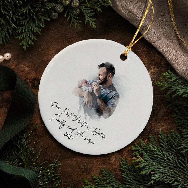 A round white ceramic ornament featuring a watercolor-style illustration of a person holding a baby. The text reads Our First Christmas Together Daddy and Aurora 2025.