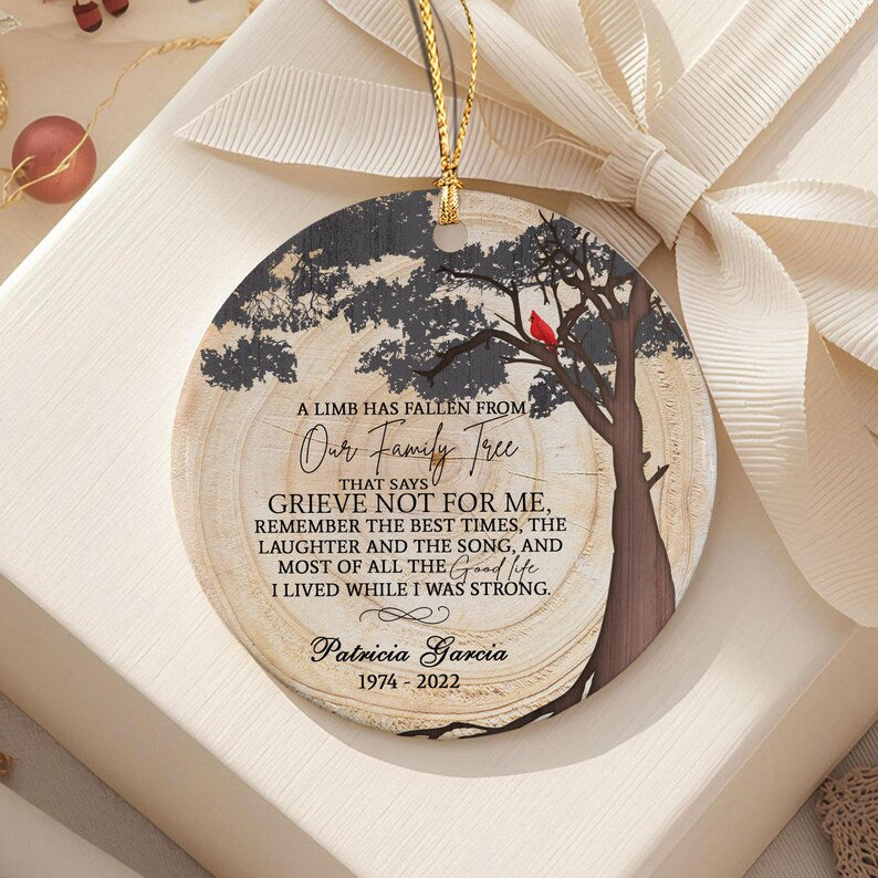 A round ceramic memorial ornament featuring a wood grain background, a dark tree silhouette with a red cardinal, and the text: A LIMB HAS FALLEN FROM OUR FAMILY TREE THAT SAYS GRIEVE NOT FOR ME, REMEMBER THE BEST TIMES.