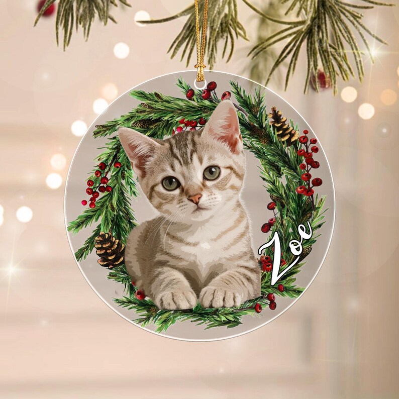 A round, flat Christmas ornament featuring a light brown tabby kitten centered within a green pine wreath decorated with red berries and pinecones. The name Zoe is printed in white script on the right side of the wreath.