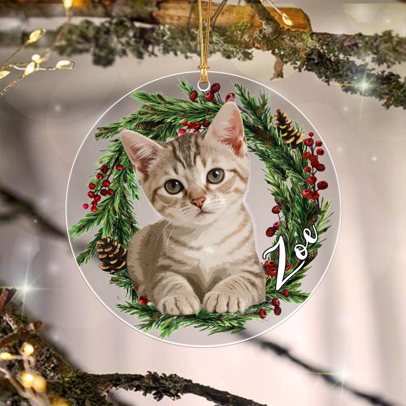 A round, flat ornament featuring a light brown tabby kitten centered within a wreath of green pine needles, red berries, and small pinecones. The name Zoe is written in white cursive script on the right side of the wreath.