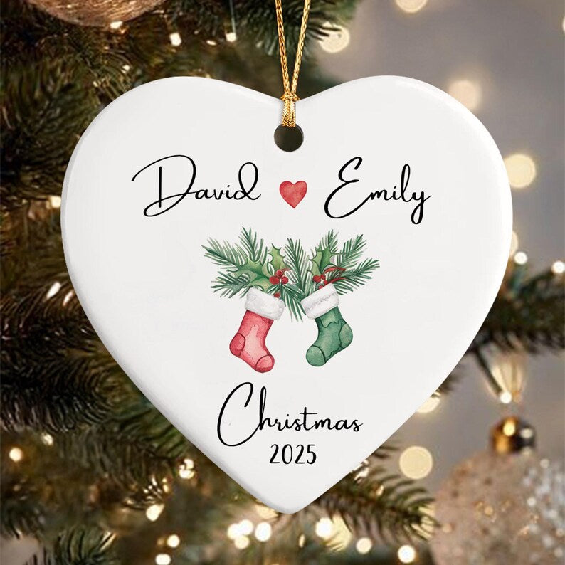 A white heart-shaped ceramic ornament featuring the names David and Emily separated by a red heart icon. Below the names is a watercolor illustration of two Christmas stockings with pine branches and the text Christmas 2025.