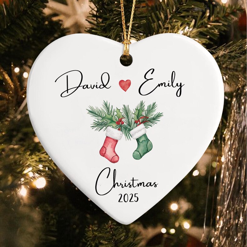 A white heart-shaped ceramic ornament featuring the names David and Emily separated by a red heart. Below, two Christmas stockings hang from green holly branches.