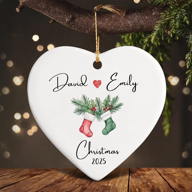 A white heart-shaped ceramic ornament featuring the names David and Emily separated by a red heart. Below, two Christmas stockings hang from pine branches, with the text Christmas 2025 printed at the bottom.