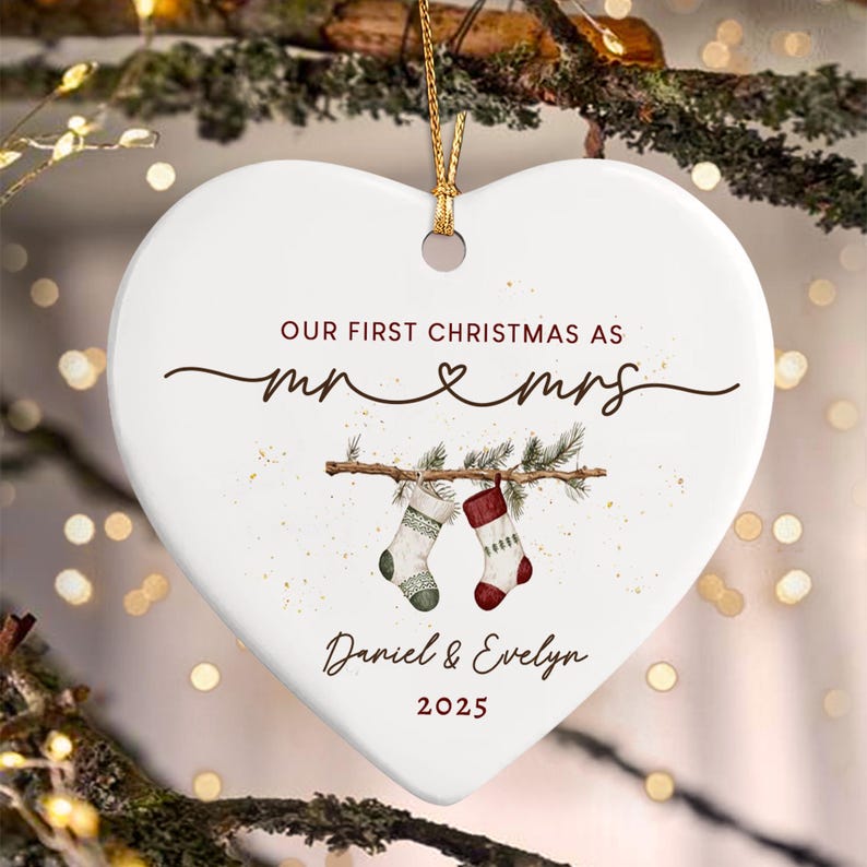 A white heart-shaped ceramic ornament featuring the text "OUR FIRST CHRISTMAS AS mr & mrs", "Daniel & Evelyn", and "2025". The design includes a graphic of two stockings hanging from a pine branch with gold string.