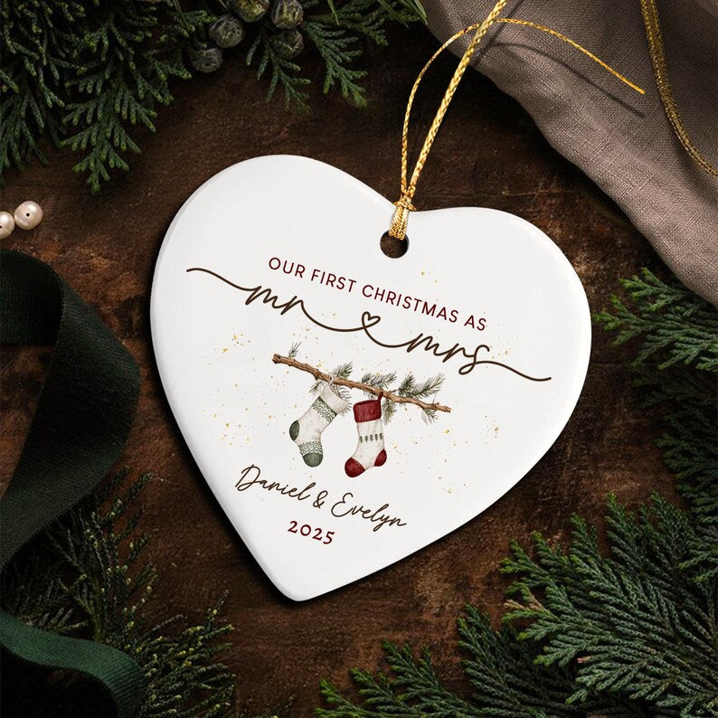 A white heart-shaped ceramic ornament with a gold hanging string. It features the text "OUR FIRST CHRISTMAS AS MR & MRS", the names "Daniel & Evelyn", and the year "2025" with an illustration of two stockings on a branch.