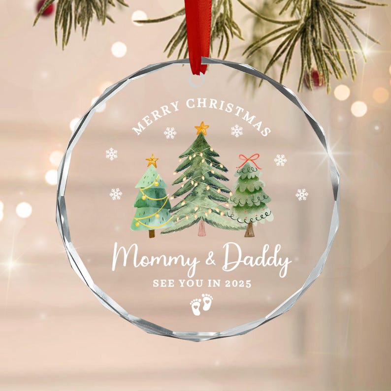 A clear, round Christmas ornament with a red ribbon. The ornament features the text "Merry Christmas" and "Mommy & Daddy SEE YOU IN 2025" with three illustrated Christmas trees and snowflakes.